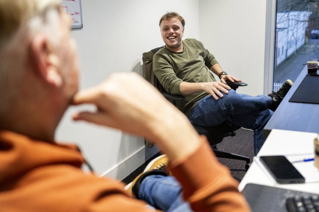 Twee collega’s in gesprek in een vergaderruimte; informele sfeer met lach en ontspannen houding.