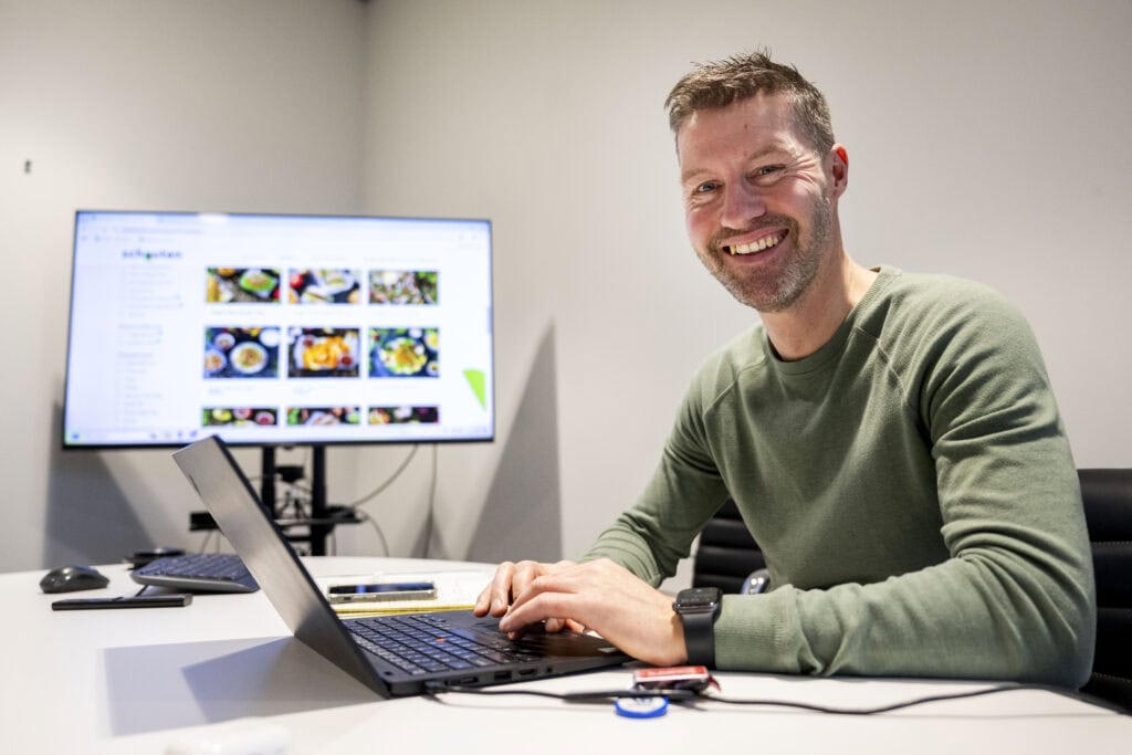 Collega lacht terwijl hij werkt achter een laptop met productfoto’s op het scherm.